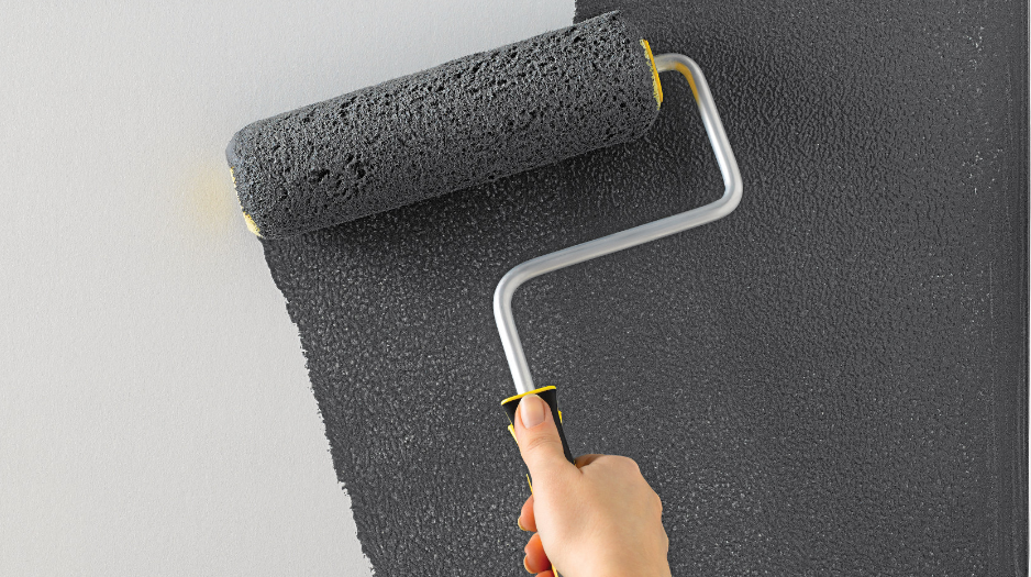 A hand applies grey paint to a white wall using a paint roller.