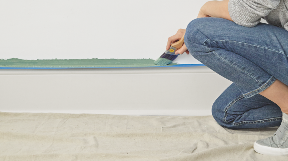 A woman kneels as she applies green paint to a wall above a baseboard using a paintbrush.