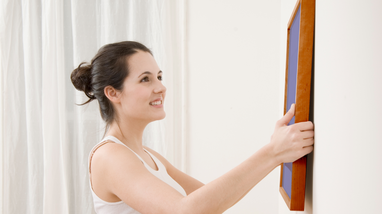 A woman happily removes a piece of framed art from a white wall in preparation of painting that wall.