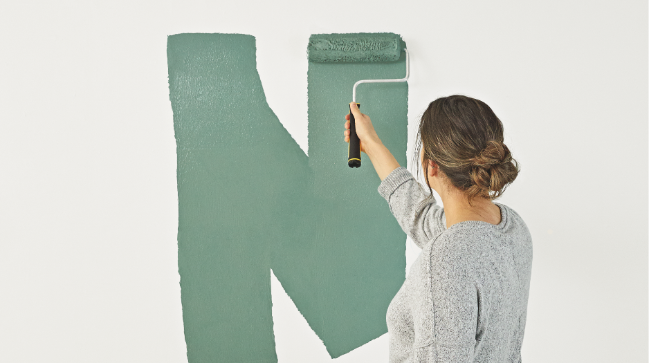 A woman applies light green paint to a white wall using a paint roller.