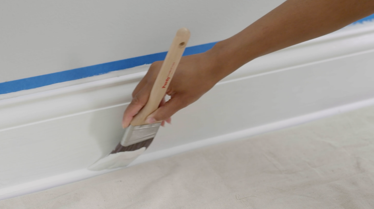 A woman’s hand applies white paint to the baseboard of an interior room using a paintbrush. Painter’s tape protects wall.