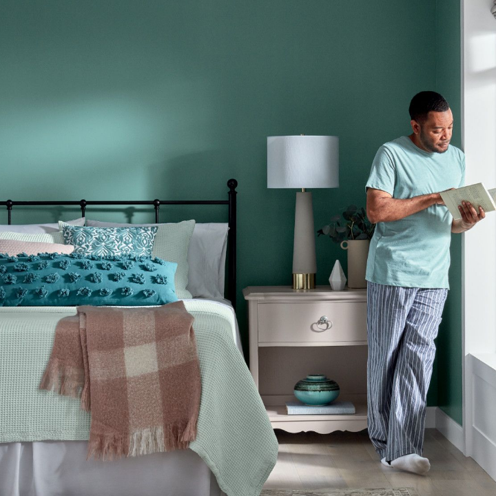 A man standing in his bedroom, leaning against a window, reading a book. Walls are painted in the color vintage teal.