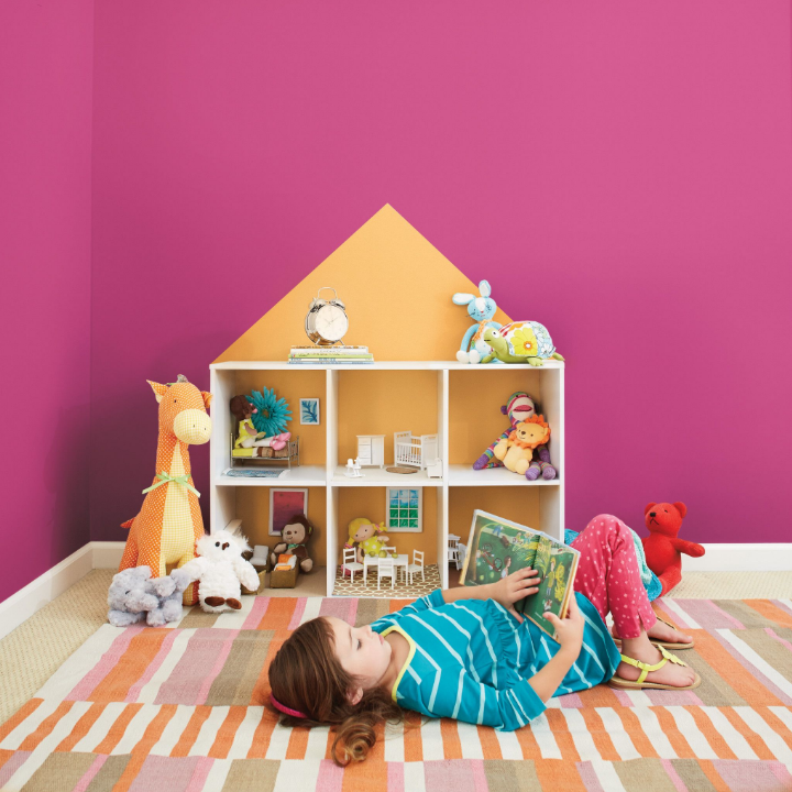 Child lays on a rug in a playroom near a doll house, reading a children’s book. Wall is painted the color glitterberry.