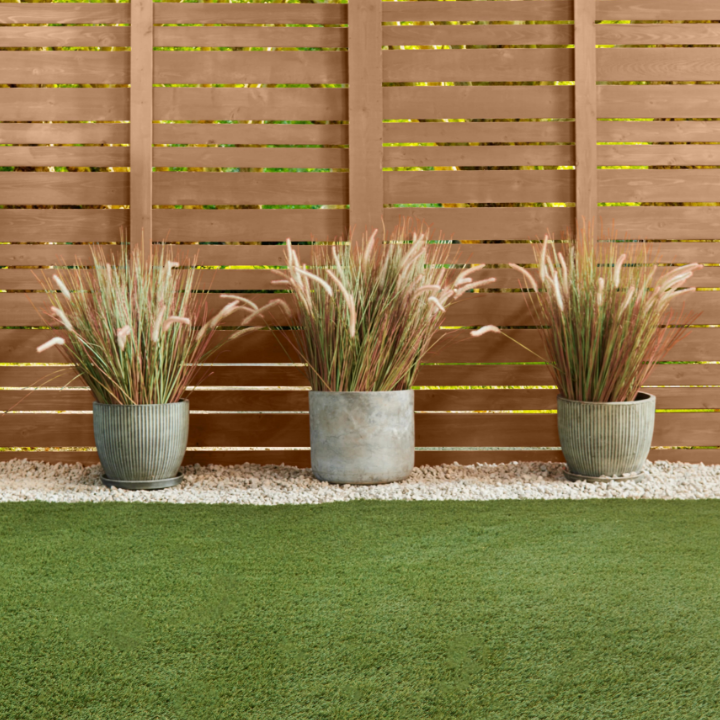 Horizontal wooden fence on side of yard, with three potted grasses over gravel in front of it. Fence painted warmed silver.
