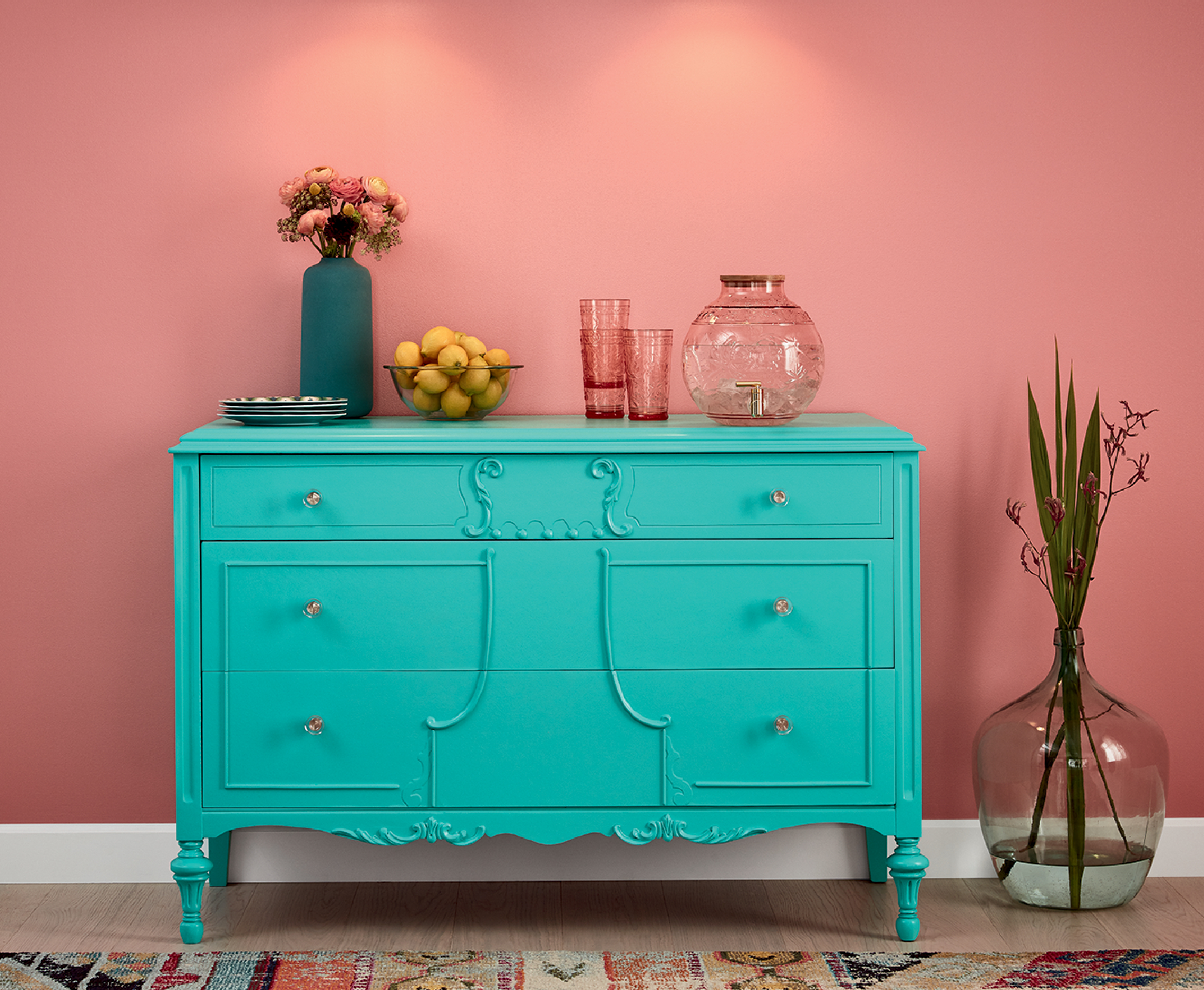 An aqua dresser sits in front of a Carmela coral wall. A vase with flowers, stack of plates and a bowl of lemons sit atop it.