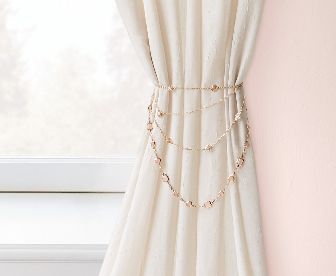 A bejeweled off-white drape sits between a window and a mauve painted wall.