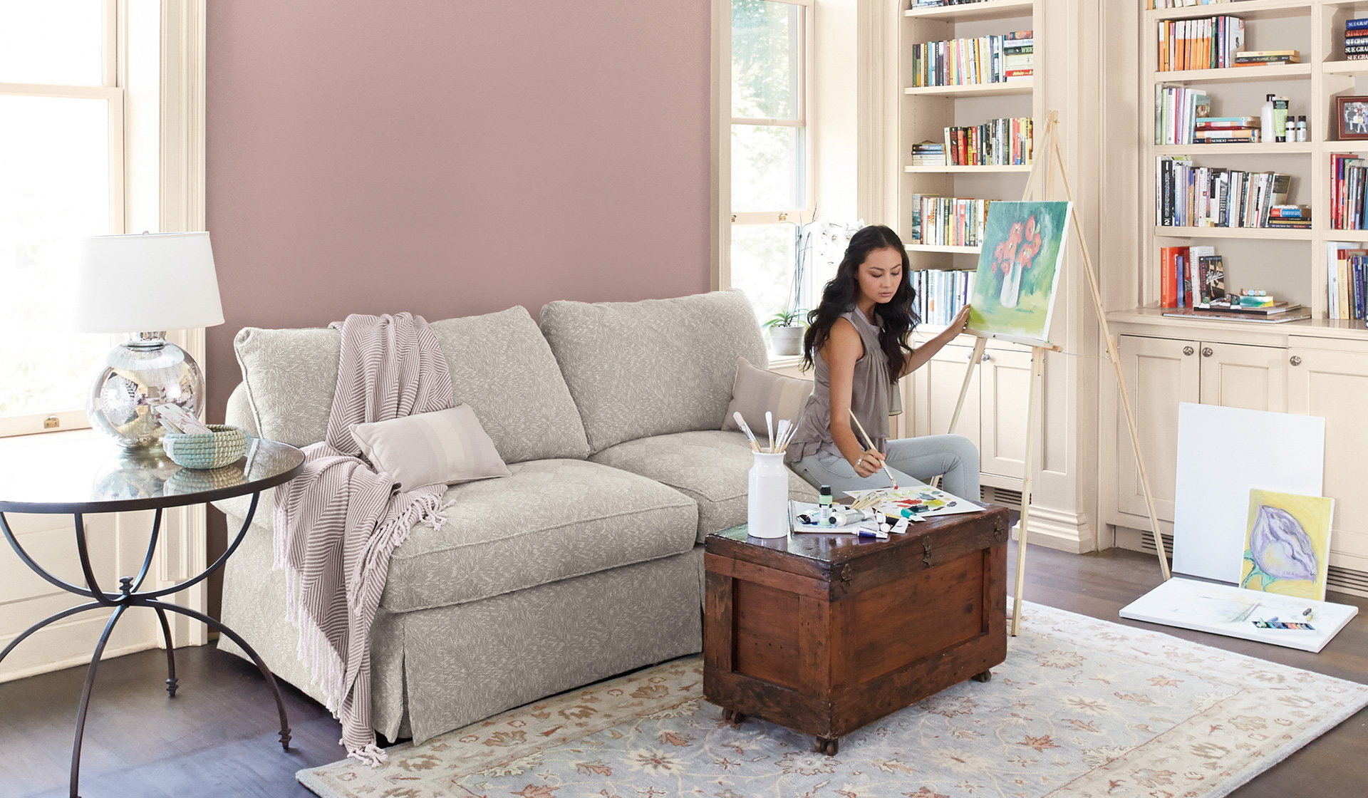A young woman sits on a couch, painting on an easel in a living room, walls painted neutral hennepin stone.