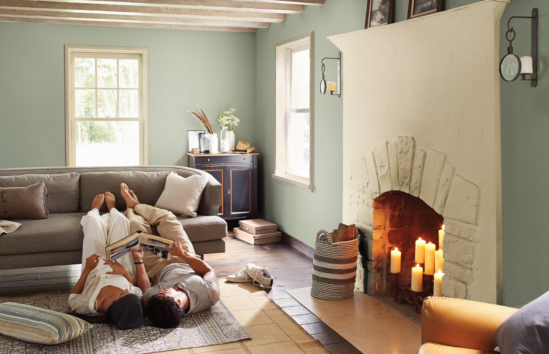 Couple lies on living room floor, feet up on couch, candles in the fireplace, reading a scrapbook. Walls painted foggy gray.