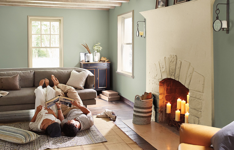 Couple lies on living room floor, feet up on couch, candles in the fireplace, reading a scrapbook. Walls painted foggy gray.