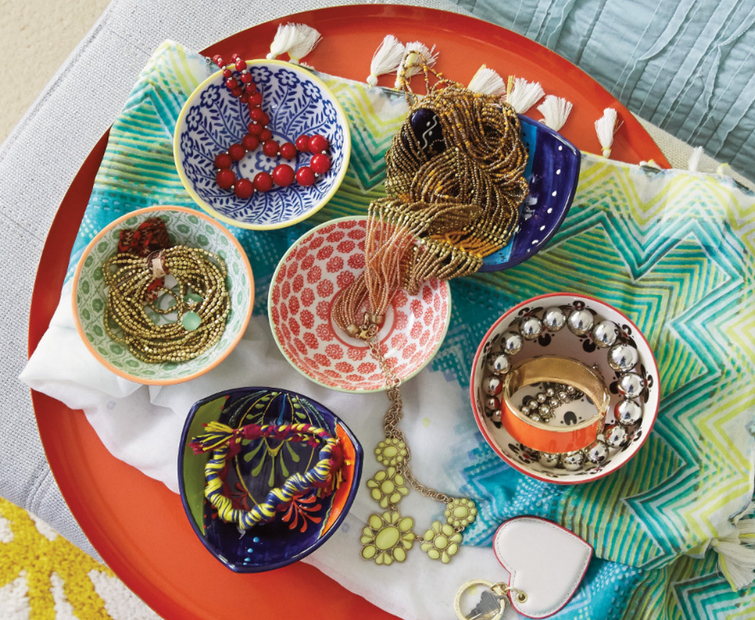Colorful bowls containing flea market jewelry sit upon a blanket and tray on a bedroom bench.