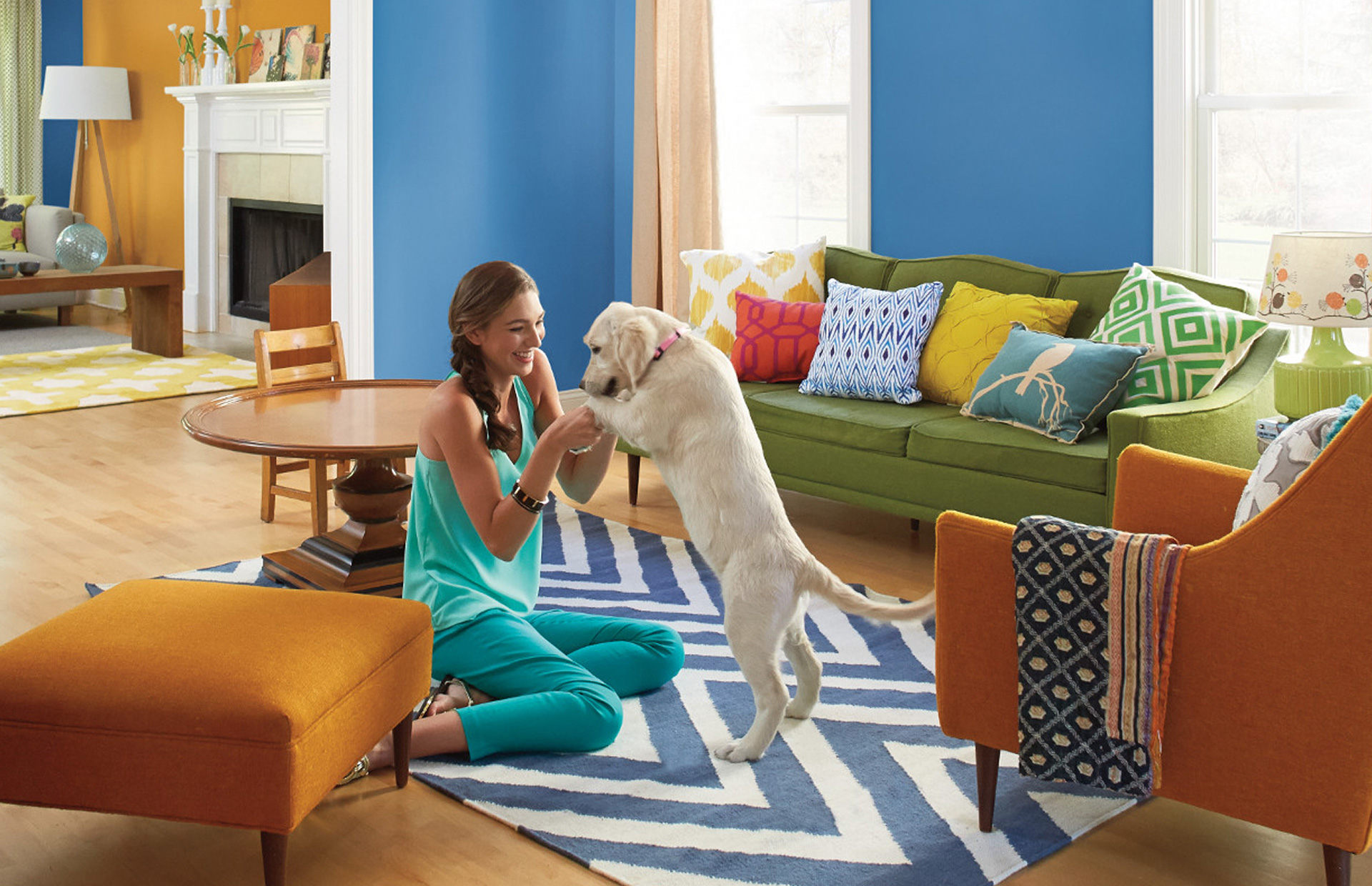 A young woman sits on a living room floor, playing happily with her white lab puppy, wall painted deep ship shade blue.