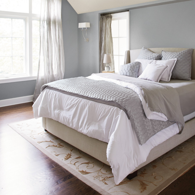 High ceiling bedroom, flooded with light from large windows. Walls painted grey with matching covers and pillows on bed.
