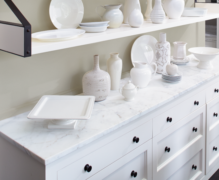A butler’s pantry with white cabinets and shelves and a marble countertop, on which sit a variety of white serving ware.