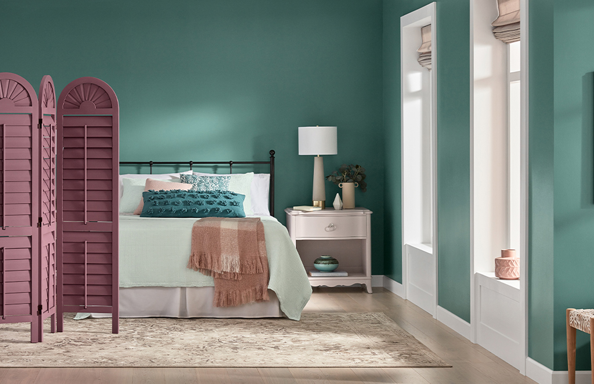 Sunlit, deep-aqua bedroom, bed with matching linens and a light pink side table and lamp, mauve temporary privacy wall.