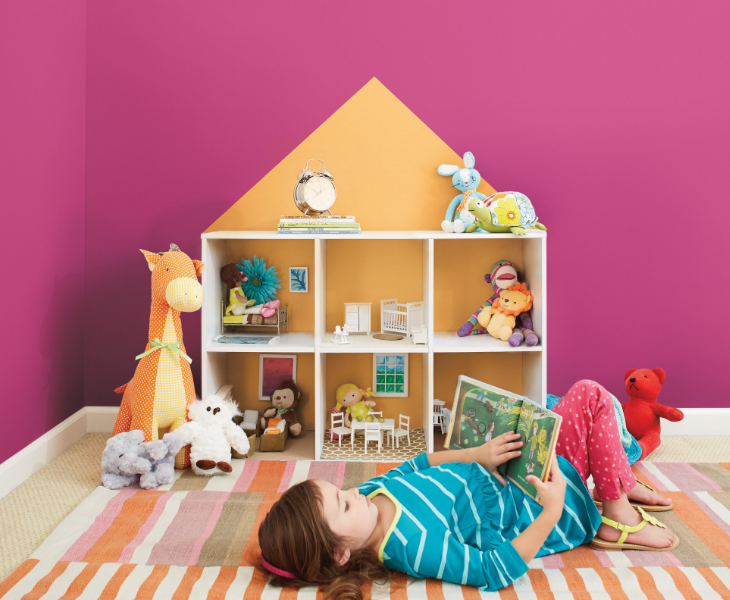 A dollhouse with stuffed animals sits in front of a deep pink wall in a lived-in kids’ bedroom.