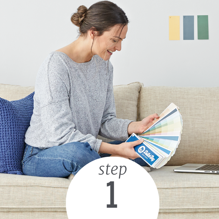 A woman sits on a couch, combing through a Dutch Boy color fan deck, choosing a one-coat color to paint.