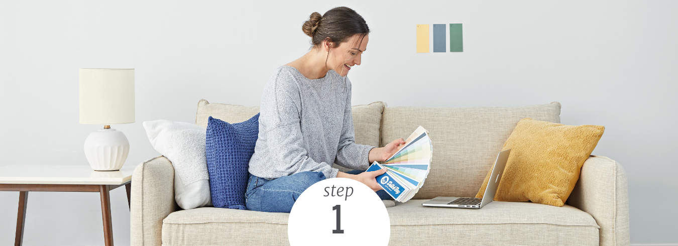 A woman sits on a couch, combing through a Dutch Boy color fan deck, choosing a one-coat color to paint.
