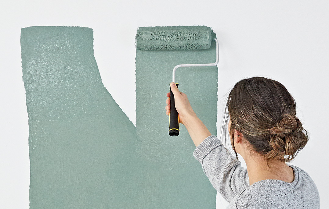 A woman stands at a wall, applying teal paint with a roller.