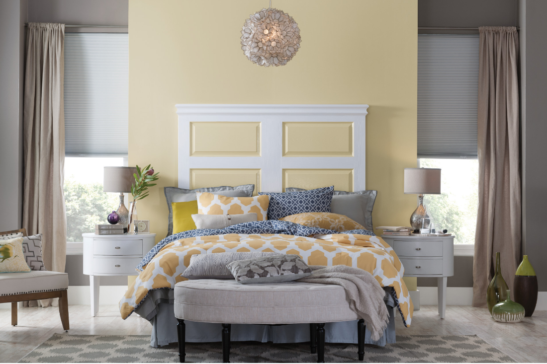 Large bedroom with yellow patterned linens and pillows paired with grey ones, wall painted yellow with matching headboard.