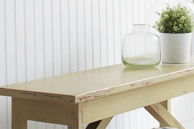 A chalky beige painted console table, with empty glass vase and potted plat atop it, sits in front of a white shiplap wall.