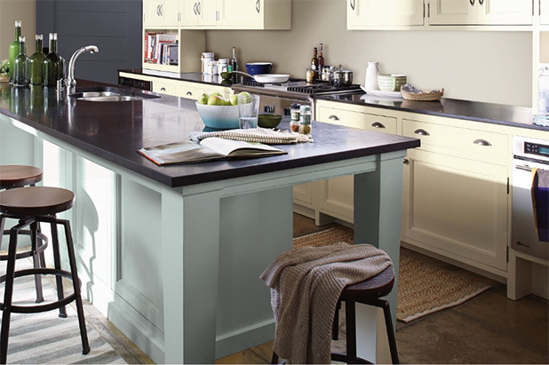 Large kitchen island with light blue cabinets in a bright kitchen that has dark stone countertops and pale-yellow cabinets. 