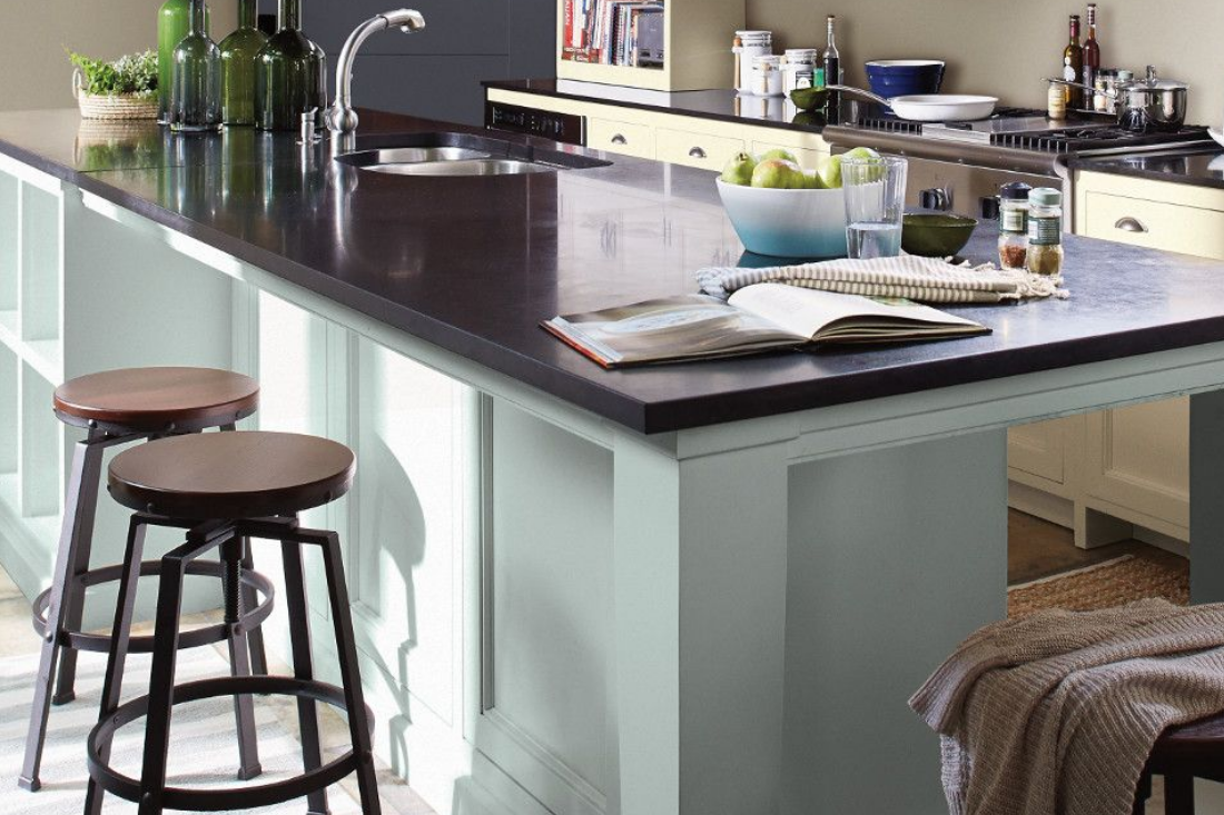 Large kitchen island with light blue cabinets in a bright kitchen that has dark stone countertops and pale-yellow cabinets. 