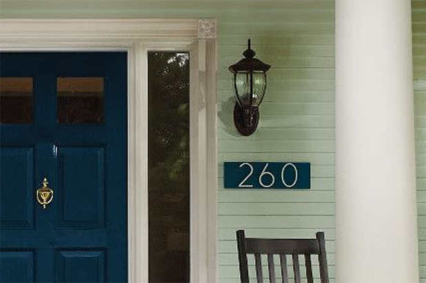 Blue front door and green siding of a house, with a rocking chair. White house address numbers on a deep sky wood backing.