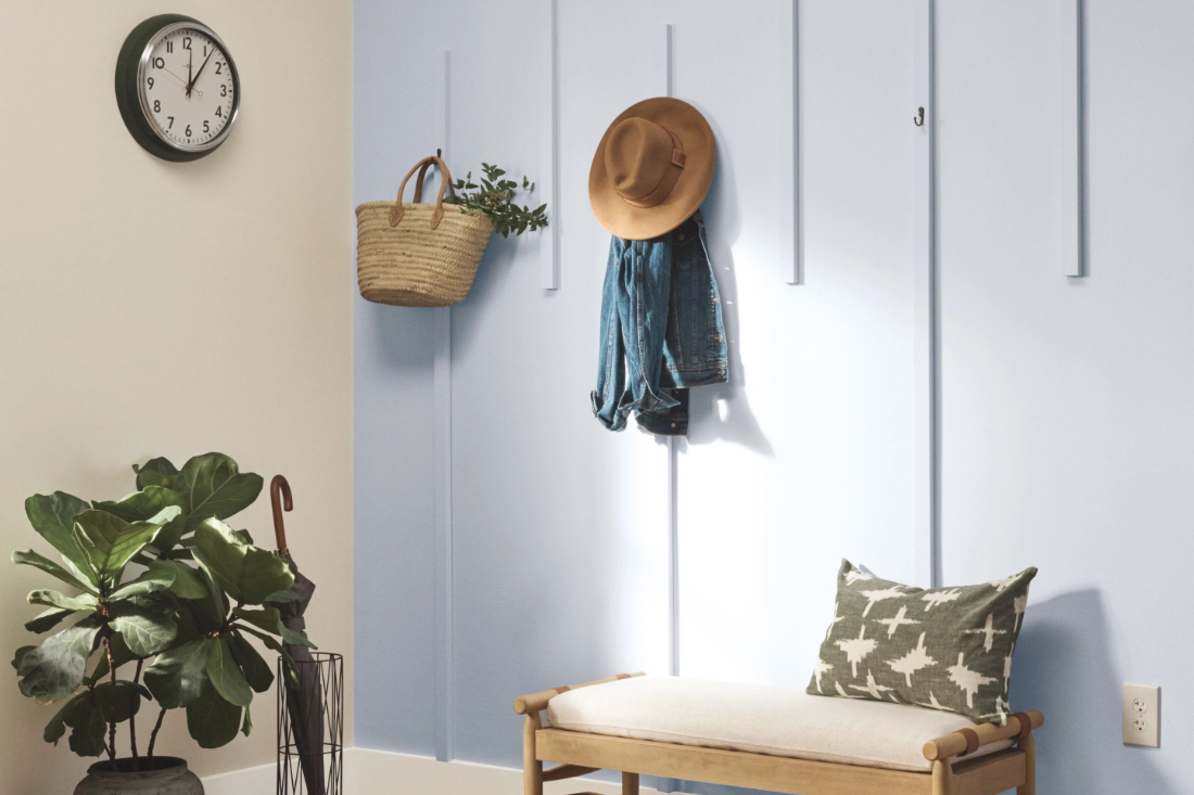 A wood bench with a pillow sits in a bright entryway, a potted plat adjacent to it.