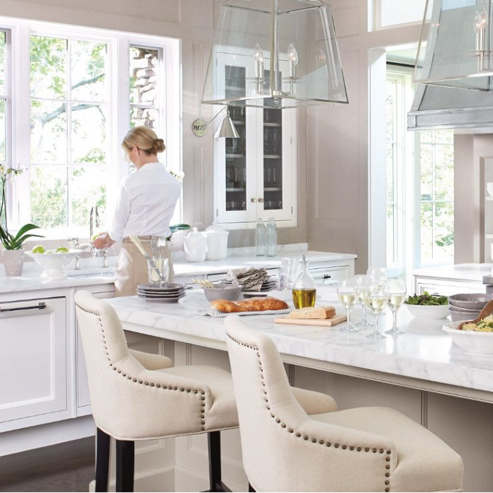Woman washes dishes in large kitchen with island, pendant lights and stools. Walls painted sultry gray.