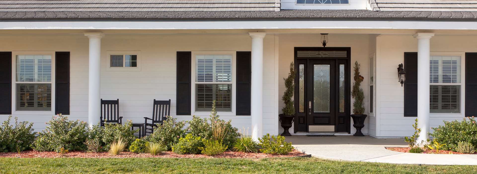 Front entrance of an upscale home with white siding and black door and shutters surrounded by beautiful landscaping