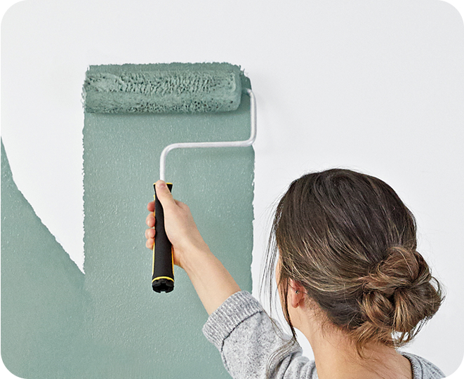 Young woman painting a wall with a roller using a sage green paint color over a white wall, showing great coverage