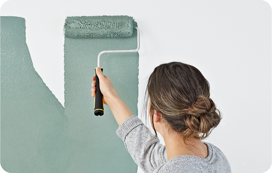 Young woman painting a wall with a roller using a sage green paint color over a white wall, showing great coverage