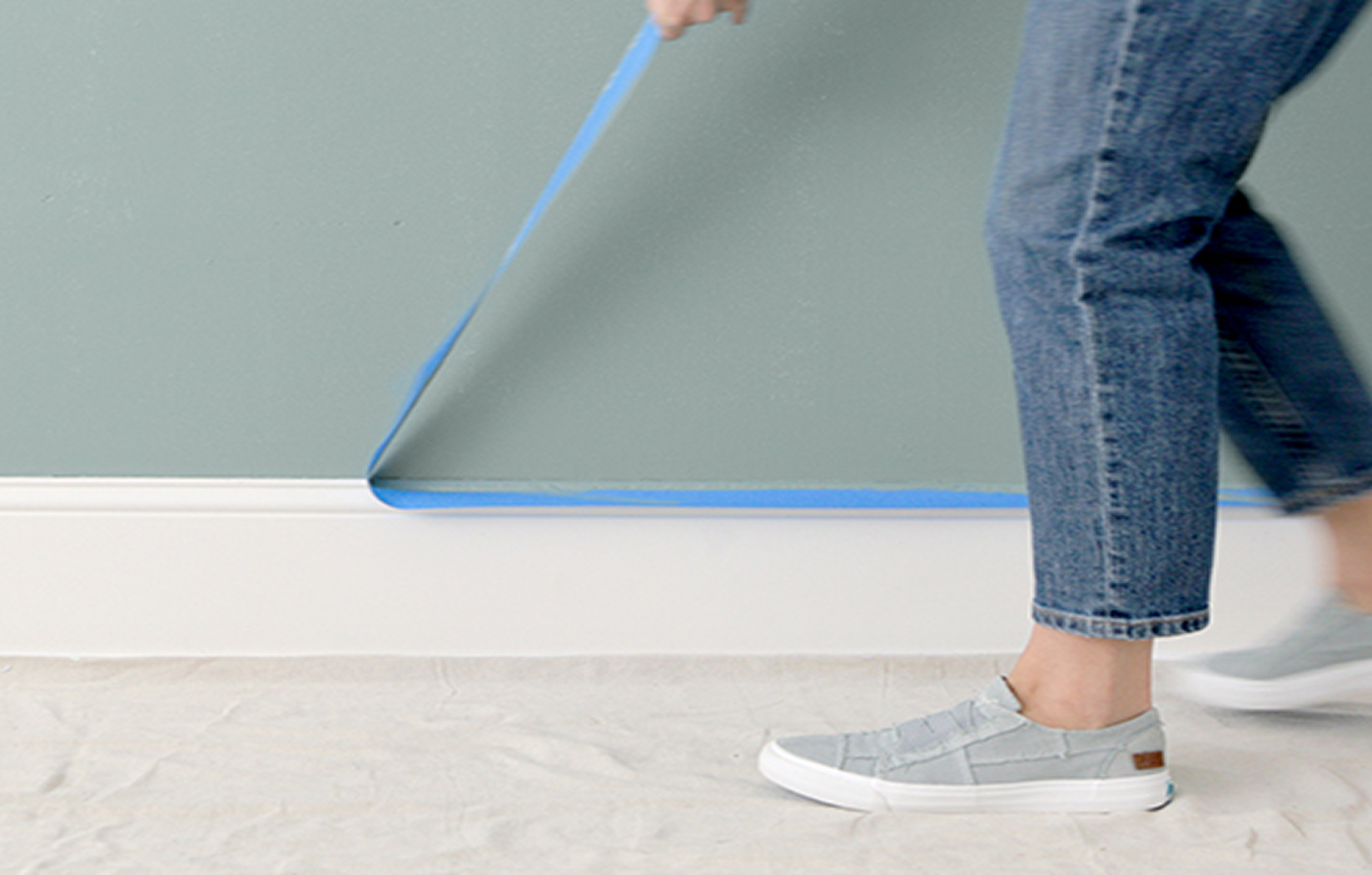 A woman walks along an interior baseboard as she removes painter’s tape from a freshly painted wall.