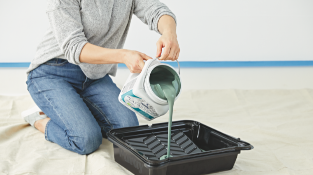 Woman kneels on floor, pouring green paint from a can into a paint tray, on top of a tarp.
