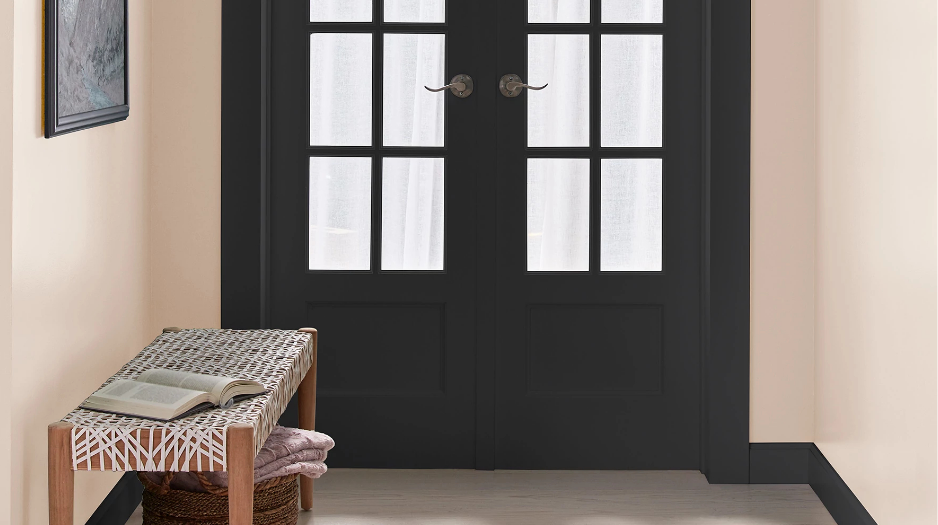 Entryway of home, French doors painted black, off-white walls, a bench with a book on it, framed art on wall.