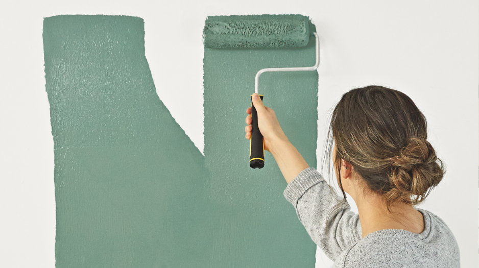 A woman applies light green paint to a white wall using a paint roller.